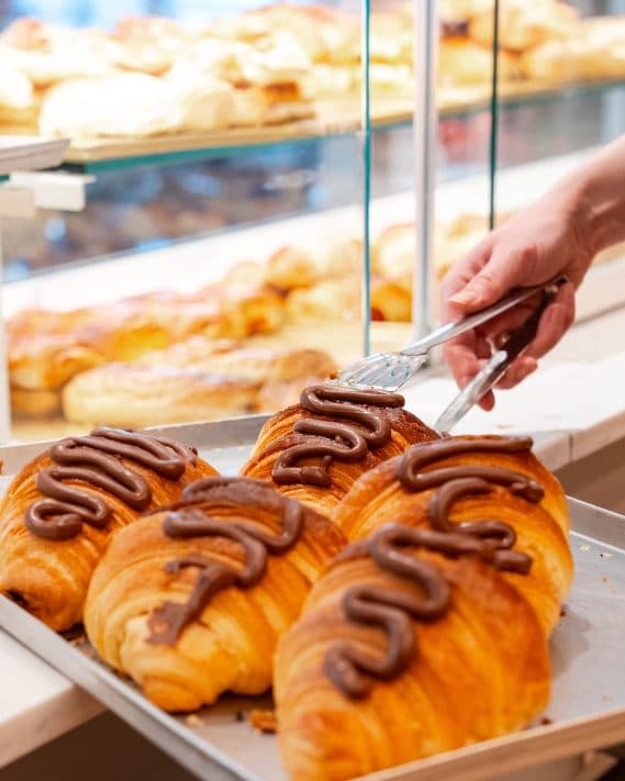 Blech mit Schokoladencroissants in der Auslage, eine Hand greift mit der Zange danach